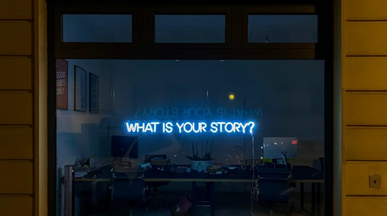 Window of a dark room with a light up sign saying "what is your story?".