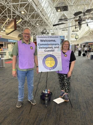 A man and a woman in light purple vests stand next to a sign that reads, "Welcome, Commissioners, Delegates and Guests!" with a GA227 logo beneath it.
