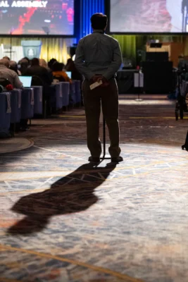 A man stands at a microphone. The photo is from behind and the man is lit up from spotlights while he faces the stage.