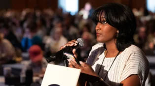 A young woman speaks that a microphone while holding her phone.