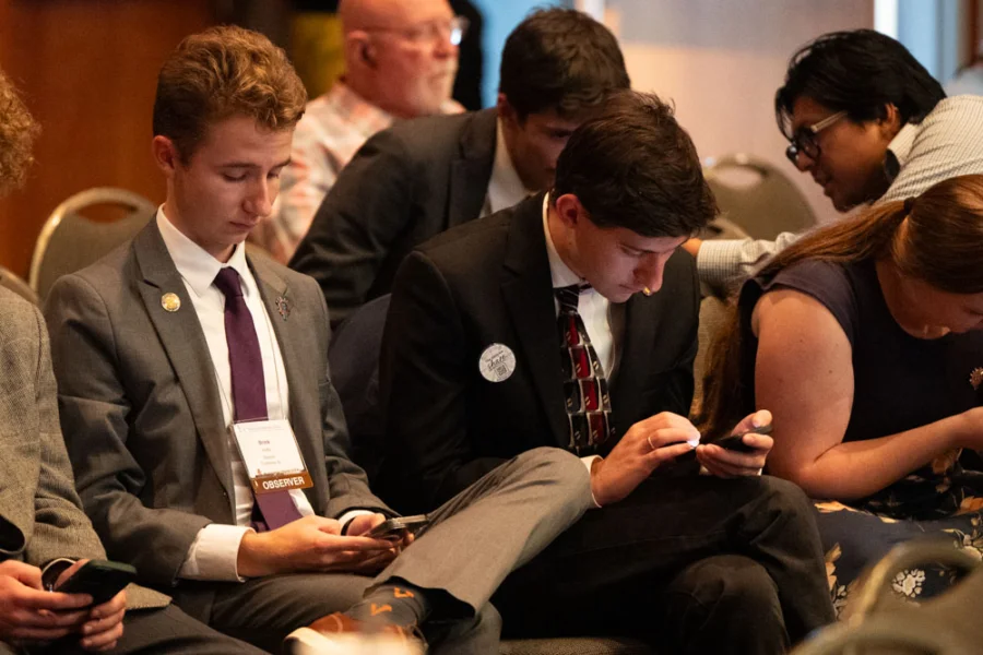 Young men in suits sit in chairs looking at their phones.