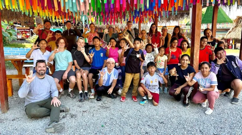 A group of children and adults pose for a picture under a pavilion with rainbow fabric hanging from the ceiling.