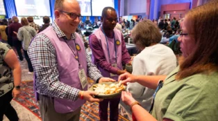Two volunteers in purple vests offer bread and grape juice for communion.