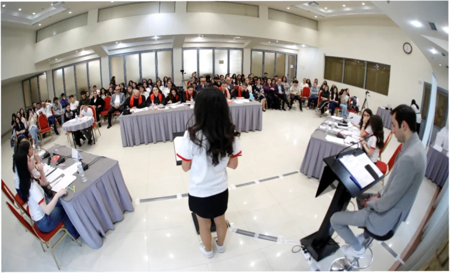 Two debate teams sit facing each other with a judges table in between them and an audience listening on. A woman stands in the middle at a microphone. The photo is taken from behind the woman who is speaking.
