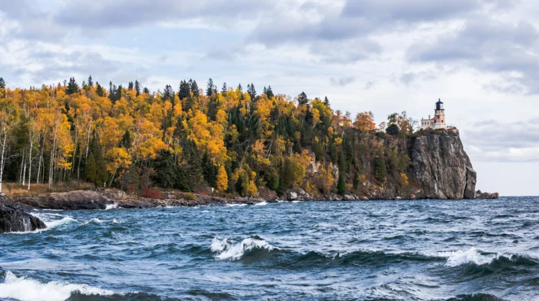 A light house on a cliff in the background. In the foreground, water and trees.