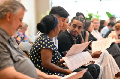 A group of young adults sit in a worship service and talk amongst themselves.