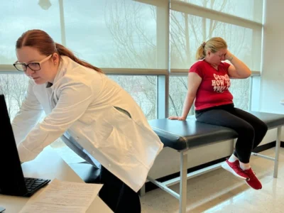Eleanor Shell sits on a medical table while a woman in a white coat types on a computer.