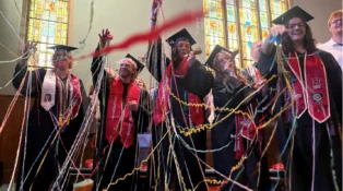 A group of college graduates from Hastings College celebrate. They are wearing their graduation garb and there are streamers falling over them.