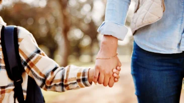 Holding hands, mom and child walking in park. Child is wearing a backpack.