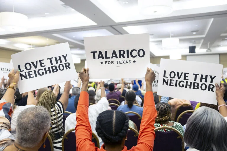 A group of people stand outside, holding signs that say, "Talarico" and "Love thy neighbor."
