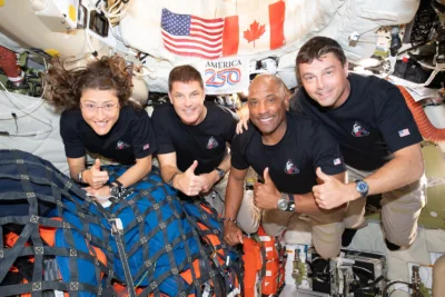 One female and three male astronauts giving thumbs up motions while floating in a spacecraft