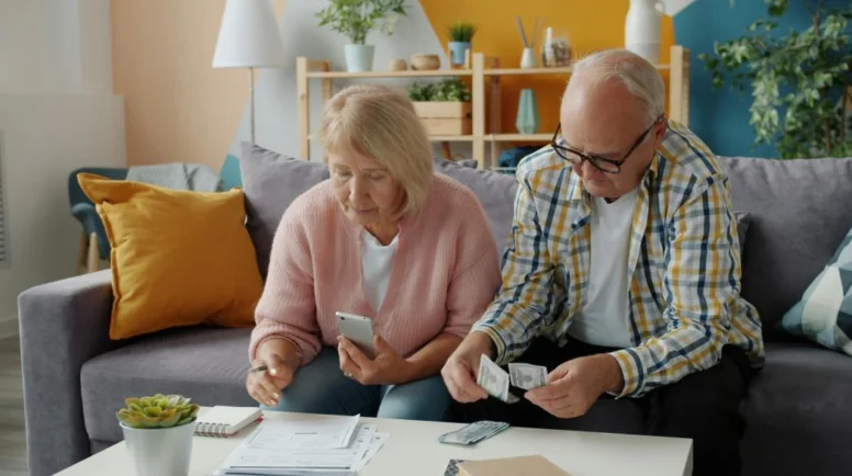 Two older adults sit on a couch with a calculator and notepad