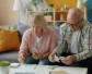 Two older adults sit on a couch with a calculator and notepad