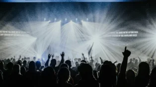 Group of people watching a worship band on stage with spotlights and smoke. People in the audience have their hands raised.
