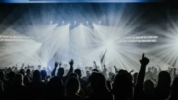 Group of people watching a worship band on stage with spotlights and smoke. People in the audience have their hands raised.