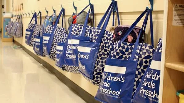 A bunch of bags hanging in a hallway. They all say "Children’s Circle Preschool"